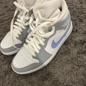 Nike Air Jordan 1 Mid grå/vit/blå - Snygga Nike Air Jordan 1 Mid sneakers i vitt och ljusgrått med en ljusblå swoosh. Klassisk high-top modell med perforerad tå, snörning och ikonisk Jordan-logga på sidan. Ovandel i skinn och syntet, perfekt för dig som vill sticka ut med stilrena färger.