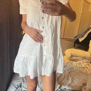 Vit kort klänning från DRYS - Säljer denna supergulliga sommarklänningen med knappar och broderier i blom mönster.  Den kommer med en underklänning så den är inte genomskinlig! Osäker på storleken tror det är en OneSize men den passar mig som är en S-M. Använd endast en gång. Inga defekter eller fläckar. Den är från märket DRYS.