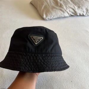 Svart bucket hat från Prada - Stilren svart bucket hat från Prada med klassisk triangellogo framtill. Hatten är tillverkad i ett lätt och slitstarkt syntetmaterial som ger en cool och modern look. Perfekt accessoar för att lyfta din outfit med en lyxig touch.