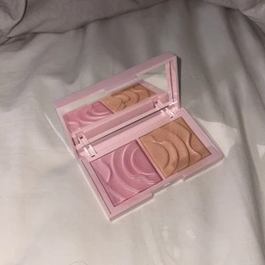 CAIA Prisma Palette Flirty highlighter - Säljer CAIA Prisma Palette Flirty, en duo highlighter i snyggt rosa fodral med spegel. Ena sidan är skimrande ljusrosa och den andra är en varm champagneguldig nyans. Båda har vågigt mönster i pudret och ger glow till ansiktet.