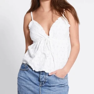 Vit broderad linnetopp med knyt - Superfin vit linnetopp med broderade blommor och smala axelband. Djup v-ringning och söt knytning framtill. Perfekt till jeans eller kjol för en fräsch sommarlook.