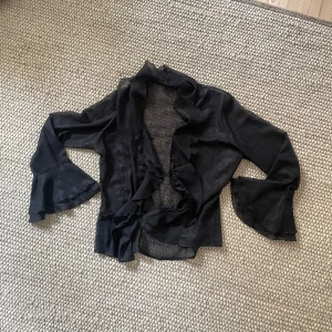 Svart volangblus - Svart transparent blus med volanger som sitter oversize på mig som är storlek S, skulle uppskatta storleken till L 🫶🏻 knappar i fram och volanger runt kragen och ärmarna 💕