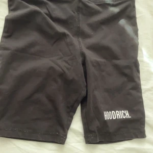 Svarta shorts från Hoodrich - Svarta, tighta shorts från Hoodrich med vit logga tryckt på ena benet. De har en enkel design och elastisk midja, perfekta för träning eller chill. Materialet känns mjukt och stretchigt, vilket gör dem bekväma att bära hela dagen.