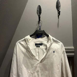 Vit slim fit skjorta Polo Ralph Lauren - Vit långärmad skjorta från Polo Ralph Lauren i slim fit-modell. Klassisk oxfordväv i bomull med stretch, broderad marinblå logga på bröstet och knappar framtill. Perfekt för dig som gillar stilrena och tidlösa plagg. Väldigt bra sick