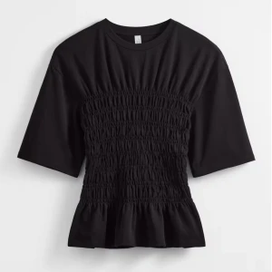 Svart smockad topp med volang - Svart topp med smockad midja och volang nertill. T-shirtrund hals, korta ärmar och en figurnära passform över magen. Perfekt för dig som gillar en enkel men ändå trendig look. Så fin använd 1 gång, privat för egna bilder❤️‍🔥 orginalpris ca 450kr