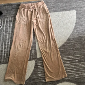 Beiga mjukisbyxor Juicy Couture - Supermjuka beiga mjukisbyxor från Juicy Couture med bred resår och snörning i midjan. Byxorna har raka ben, stora fickor fram och bakfickor med lock och knapp. Perfekta för en chill och avslappnad stil.