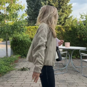Grön bomberjacka i linne  - Snygg grön bomberjacka med rund halsringning och dragkedja framtill. Jackan har två fickor med knappdetaljer och elastisk nederkant för en chill passform. 