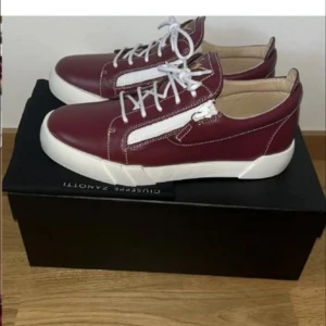 Giuseppe Zanotti - Snygga vinröda sneakers från Giuseppe Zanotti i skinn med vita snören och dragkedjor på sidorna. Skorna har vit platt sula och rund tå, samt guldfärgad logga på plösen. Perfekta för dig som vill sticka ut med stilrena detaljer.