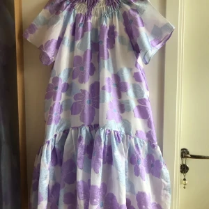 Ljuslila blommig volangklänning - Superfin volangklänning med stora lila och ljusblå blommor på vit botten. Klänningen har korta ärmar, smockad halsringning och luftig passform. Perfekt för dig som gillar romantiska och färgglada plagg med extra volym.