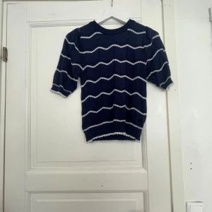Blå stickad tröja Holly & Whyte S - Mörkblå stickad tröja från Holly & Whyte med vita vågiga ränder. Tröjan har korta puffärmar och rund halsringning. Snygga detaljer med ribbade muddar och liten volangkant vid ärmslut och nederkant. Perfekt för dig som gillar en preppy och söt stil.