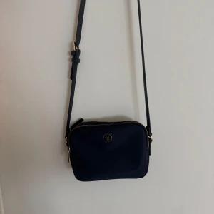Mörkblå axelväska från Tommy Hilfiger - Snygg mörkblå axelväska från Tommy Hilfiger med justerbar rem och guldfärgade detaljer. Väskan har två fack med dragkedja och klassisk TH-logga framtill. Materialet är slätt och stilrent, perfekt för att bära dina essentials.
