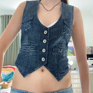 Jeansväst från NINELIVES Anarchy - Snygg jeansväst i mörkblå denim från NINELIVES Anarchy. Fyra fickor framtill, markerad midja och cool spetsig nederkant. Fyra silvriga knappar och har en djup v-ringning. Perfekt att styla över en t-shirt eller linne till sommaren eller hösten. 📏Ungefära mått(hela vägen runt): byst-70cm midja-65cm