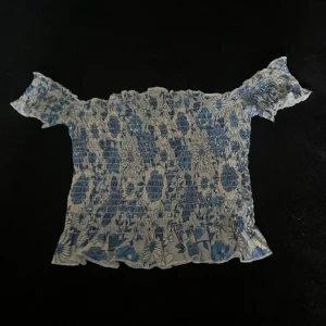 Blommig offshoulder topp från Bik Bok - Supersöt offshoulder topp från Bik Bok i vitt och blått med blommigt mönster. Smockad över hela toppen och volangkant nertill och vid ärmslut. Perfekt för dig som gillar romantisk och somrig stil.