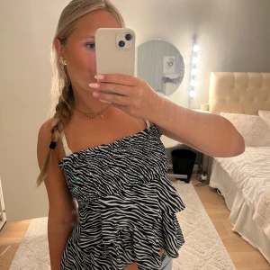 Zebramönstrad volangtopp från Gina Tricot - Supersöt zebramönstrad topp från Gina Tricot med volanger och smock upptill. Toppen är ärmlös och har en luftig, flowy passform som känns riktigt trendig. Perfekt för dig som vill sticka ut med ett djurmönster och coola detaljer.