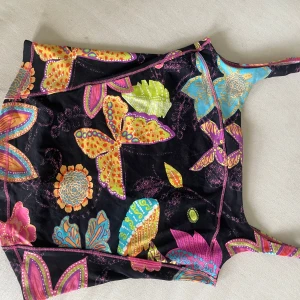Färgglad linnetopp från Fuego Woman - Superfin linnetopp från Fuego Woman med färgstarkt blommigt och fjärilsmönster i gult, orange, blått, rosa och grönt mot svart bakgrund. Toppens smala axelband och mjuka material gör den perfekt för varma dagar. Snygga kontrastsömmar i rosa längs kanterna.