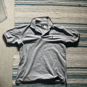 Grå piké t-shirt med rosa detaljer - Snygg grå piké från Polo Ralph Lauren med klassisk krage och rosa kantdetaljer. T-shirten har korta ärmar och liten broderad logga på bröstet. Perfekt för en clean och stilren look.