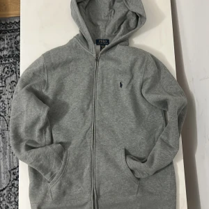Grå hoodie från Polo Ralph Lauren - Klassisk grå hoodie från Polo Ralph Lauren med dragkedja och huva. Tröjan har broderad logga på bröstet, två fickor fram och är tillverkad i mjuk bomullsmix. 