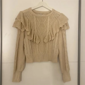 Beige stickad tröja med volang - Superfin beige stickad tröja med volangdetaljer över axlarna och kabelstickat mönster. Tröjan har lång ärm och ribbade muddar vid ärmslut och nederkant. Perfekt för dig som gillar en söt och trendig look.