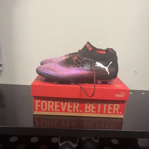 Puma Future Z 1.3 fotbollsskor strl 44 - Säljer ett par Puma Future Z 1.3 fotbollsskor i storlek 44. Skorna har en svart bas med lila och röda detaljer samt vita Puma-loggor på sidorna. Ovandelen är i syntetmaterial med snörning och en strumpliknande passform för extra stöd på planen. Sulor finns om man önskar