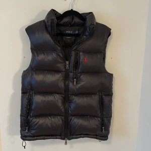 Svart puffer dunväst från Polo Ralph Lauren - Snygg svart dunväst från Polo Ralph Lauren med klassisk röd logga broderad på bröstet. Västen har hög krage, två dragkedjefickor och quiltad design. Yttermaterialet är glansigt och tillverkat i återvunnen nylon. Perfekt att slänga över en tjocktröja