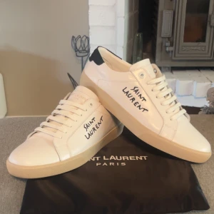 Saint Laurent vita sneakers i skinn - Snygga vita sneakers från Saint Laurent med svart detalj på hälen och handskriven logga på sidan. Skorna har klassisk rund tå, snörning och platt sula i beige. Tillverkade i slätt skinn för en clean och lyxig look. Box medföljer🤗