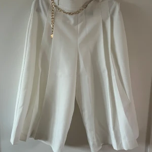 Vit plisserad byxkjol med kedjedetalj - Snygg vit knälång  byxkjol  med breda plisserade veck och en guldig kedjedetalj i midjan. Kjolen har en elegant och flowy siluett som ger en cool och trendig vibe. Perfekt för dig som vill sticka ut med en stilren look.