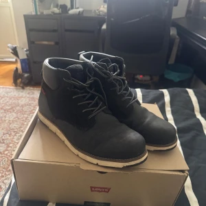 Svarta boots från Levi's med snörning - Snygga svarta boots från Levi's med robust vit sula och klassisk snörning. Skorna har en rund tå och är tillverkade i skinn med detaljer i mocka. Perfekta för höst och vinter när du vill ha både stil och komfort.