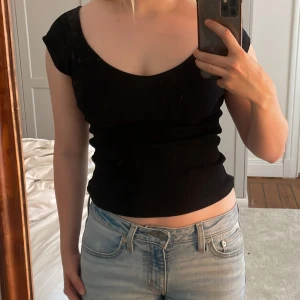 Svart ribbad topp med kort ärm - En svart, tight ribbad topp från BSL med korta ärmar och rundad urringning. Sitter snyggt figurnära och är perfekt till jeans eller kjol. Enkel och stilren design som funkar till många olika looks.