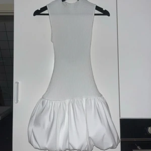 Vit kort klänning från Zara - Superfin vit kort klänning från Zara med ribbad överdel och voluminös ballongkjol. Ärmlös modell med rund hals och stor öppen rygg som ger en cool och modern look. Perfekt för dig som vill sticka ut med en unik siluett.