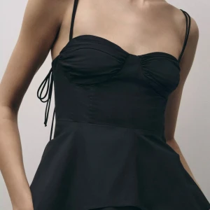 Svart volangtopp med knytband från Zara - Helt oanvänd!! Supersnygg svart korsettopp med tunna axelband och volang nertill. Toppen har rynkade kupor, smock i ryggen och knytband i sidorna för extra detalj. Perfekt för dig som vill ha en trendig och feminin look.