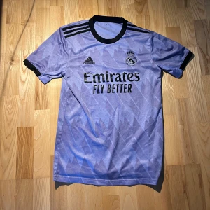 Real Madrid Adidas fotbollströja lila - Snygg Real Madrid fotbollströja från Adidas i ljuslila med mörka detaljer och geometriskt mönster. Tröjan har svarta ränder på axlarna, svart krage och klubbmärke samt sponsortryck på bröstet. Perfekt för dig som älskar fotboll och vill sticka ut på planen.
