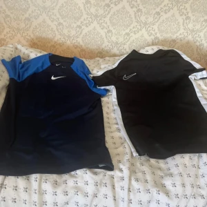 Nike Dri-Fit fotbollströjor blå & svart - Två Nike Dri-Fit fotbollströjor säljes! En är marinblå med blå axlar och vit Nike-logga, den andra är svart med vita sidor och vit logga. Båda är kortärmade och tillverkade i funktionsmaterial som andas. Perfekta för träning eller match! S och M storlek 