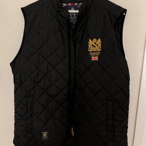 Svart quiltad väst från Morris - Snygg svart quiltad väst från Morris med guldbroderad logga och brittisk flagga på bröstet. Västen har dragkedja framtill och två fickor. Perfekt för lager-på-lager och stilren look.