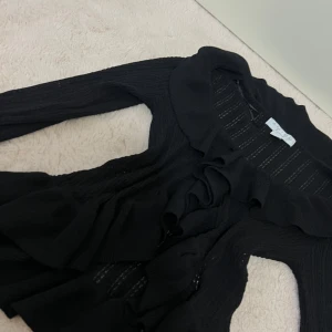 Svart volangkofta från Amisu,  - Svart stickad kofta från Amisu i storlek L. Koftan har långa ärmar, volangkant längs öppningen och ett lätt genombrutet mönster i tyget. Perfekt för lager-på-lager och ger en chic vibe till din outfit. Den är lite sliten men det går att sy upp! Passar xs s m l