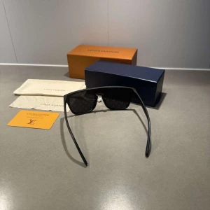 Svarta solglasögon Louis Vuitton - Snygga svarta solglasögon från Louis Vuitton med bred, rak båge och mörka glas med diskret LV-monogram. Glasögonen har en modern, kantig form och är tillverkade i ett lätt syntetmaterial. Perfekt accessoar för dig som vill sticka ut med exklusiv stil.