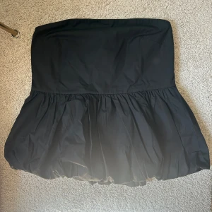 Svart tubtopp från Kappahl - Supersöt svart tubtopp från Kappahl med   ballongformad nederdel. Toppen är axellös och har smock baktill för extra bekväm passform. Perfekt att styla med ett par jeans. Helt ny med lapp kvar. .