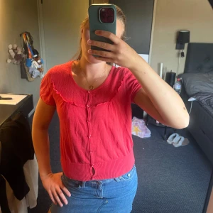 Röd blus med volang och knappar - Supersöt rosa blus med korta ärmar, volangdetalj längs kragen och knappar hela vägen framtill. Ribbad nederkant ger en snygg passform. 