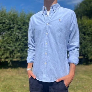 Ljusblå skjorta från Ralph Lauren - Snygg ljusblå skjorta från Ralph Lauren i slim fit-modell. Skjortan är i 100% bomull, har klassisk krage, långa ärmar och knäppning framtill. Ikoniska broderade polospelaren på bröstet ger en clean och stilren look.