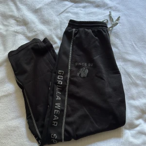 Svarta mjukisbyxor Gorilla Wear L/XL - Svarta mjukisbyxor från Gorilla Wear med tryckt logga och text längs benen. Byxorna har resår och snörning i midjan samt kontrastsömmar. Perfekta för gymmet eller chill, tillverkade i mjukt syntetmaterial för maximal komfort.