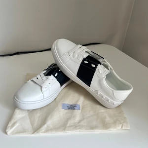 Valentino Garavani Open sneakers ÄKTA‼️ - Snygga Valentino Garavani Open sneakers i vitt skinn med svart bred rem över mitten. Klassisk rund tå, vita snören och ikoniska nitar på hälen. Perfekta för dig som vill ha en clean men ändå lyxig look. OBS ÄKTA‼️Köpt i butik i Italien på påsklovet och endast använda ett fåtal gånger bl.a skolavslutningen men nu står dom och samlar damm på rummet. Kan gå ner i pris vid snabb affär, byten är intressant men inget skambud😁