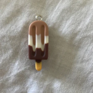 Glasspinne-halsbandsberlock i brunt. - Supergullig berlock formad som en glasspinne i bruna, vita och gula nyanser. Perfekt att hänga på ett halsband för en lekfull och somrig vibe. Tillverkad i plast med en liten metallögla upptill för enkel fastsättning.