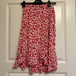 Röd vit blommig knyt omlottkjol Indiska - Supersöt röd omlottkjol med vita blommor och volangkant. Kjolen har en flowy känsla och knyts i sidan för en snygg passform. Perfekt för varma dagar. Inköpt på Indiska. Aldrig använd.