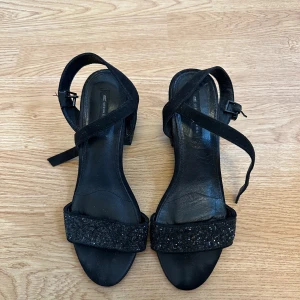 Svarta glittriga sandaler sandaletter med klack - Svarta sandaler från Urban Classics med glitter-detaljer. Skorna har öppen tå, justerbar rem runt ankeln och en stadig klack. Perfekta för dig som vill sticka ut med lite extra glitter.