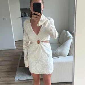 Vit kortklänning med cut out och ring - Superfin vit kortklänning med lång ärm, djup v-ringning och snygg cut out i midjan med en dekorativ ringdetalj framtill. Klänningen har ett diskret blommigt mönster i tyget och är perfekt för sommarens alla tillfällen.