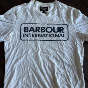 Vit Barbour International t-shirt - Vit t-shirt från Barbour International med svart logga tryckt på bröstet. Klassisk rund hals och korta ärmar. Modellen är slim och i mjuk bomull, perfekt för en clean och stilren look.