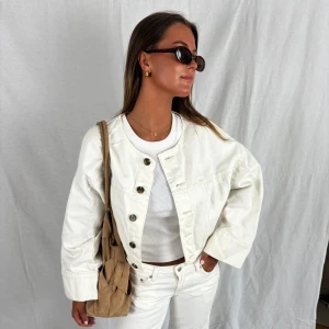 Vit croppad jeansjacka med knappar - Säljer en vit croppad oversized jeansjacka med stora bruna knappar och rund halsringning. Jackan har en loose passform och långa ärmar, perfekt att styla med matchande jeans eller kjol. Enkel och clean look som passar till många outfits.