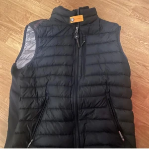 Svart dunväst från Parajumpers - Snygg svart dunväst från Parajumpers med quiltad design och hög krage. Västen har dragkedja framtill, två sidofickor med dragkedja och en gul detalj vid halsen. Insidan är ljusgrå och västen är perfekt för lager-på-lager under kyliga dagar.