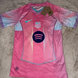 Barcelona x Nike fotbollströja  - Säljer en FC Barcelona x Nike fotbollströja i rosa och ljusblått med coola grafiska linjer och Spotify-logga på bröstet. Tröjan är kortärmad, har V-ringning med turkos detalj och är gjord i lätt, ventilerande Dri-FIT-material. Perfekt för dig som vill sticka ut på planen. Superbra för dig som vill gå runt med självförtroende och stil.