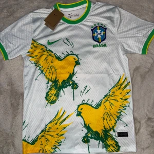 Brasilien x nike forbollströja  - Snygg vit Brasilien fotbollströja från Nike med gröna och gula detaljer på krage och ärmslut. Framsidan har stora gula fåglar i grafisk stil och CBF-emblemet broderat på bröstet. Tillverkad i Dri-FIT-material som andas, perfekt för match eller träning. Perfekt för en stilig look 