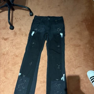 Svarta flared jeans med vita färg detaljer  - Unika svarta jeans med breda ben och vita färgstänk samt råa, ofållade kanter längst ner. Byxorna har paneler i olika svarta nyanser och en cool, konstnärlig look. Perfekta för dig som vill sticka ut med din stil.